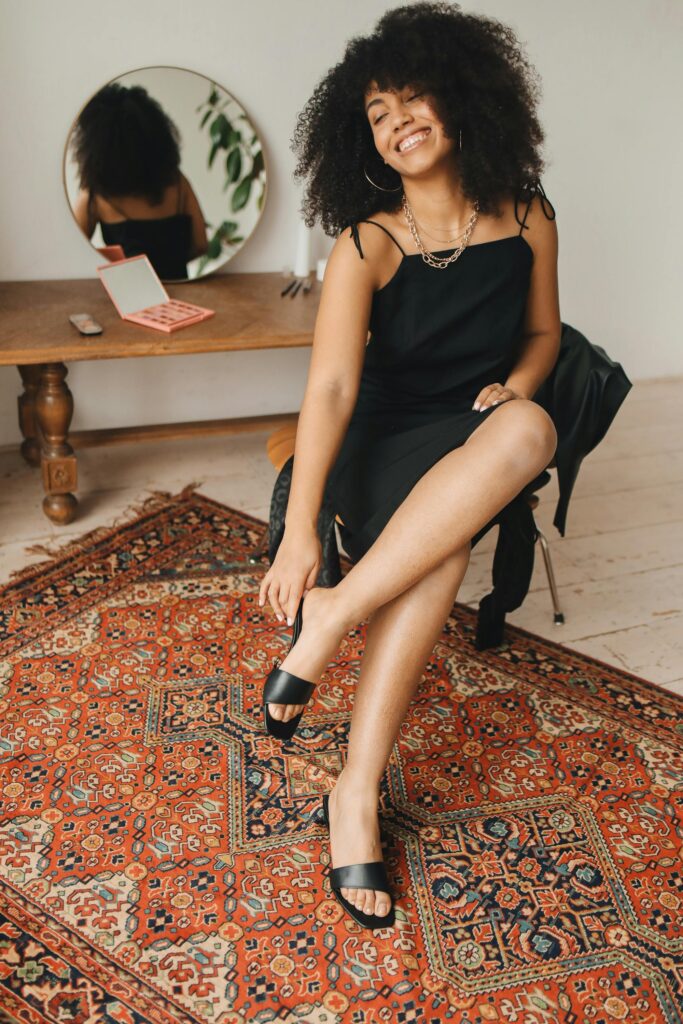 Fashionable woman in black dress smiling indoors, sitting near a mirror.